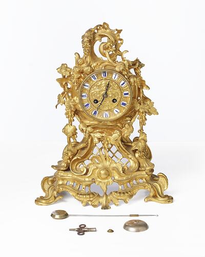 Elaborately decorated gold mantel clock. Gold face, white around roman numerals. Parts laid out in front.