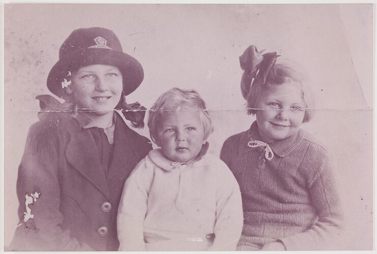 Three children pose. Eldest girl on left wears a hat, youngest boy in centre and girl on right has a bow in her hair.
