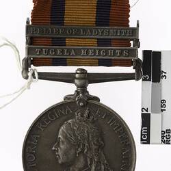 Round silver coloured medal with profile of crowned woman, text surrounding and multicoloured ribbon.