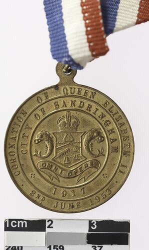 Round medal with coat of arms and text surrounding, with red, white and blue ribbon attached.