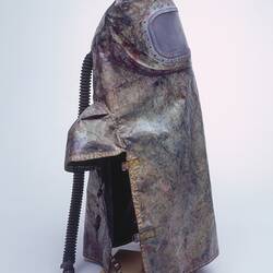 Discloured brown safety hood and apron with clear plastic section over face. Capped shoulders. Tube at back.