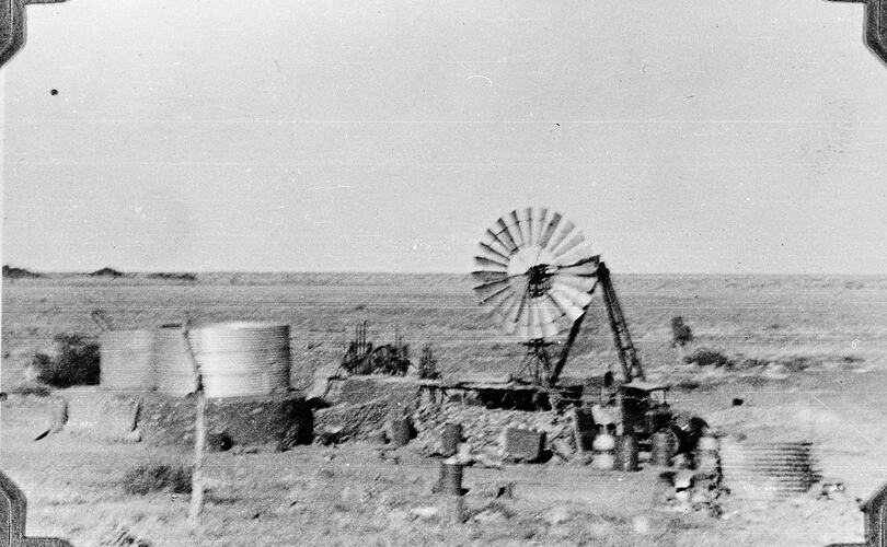 Flat barren land with truck and windmill. Two water tanks to the left.