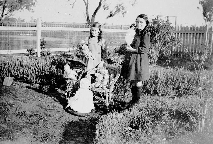 Two girls with their dolls in the garden of a house. Picket and emu wire woven fence behind.
