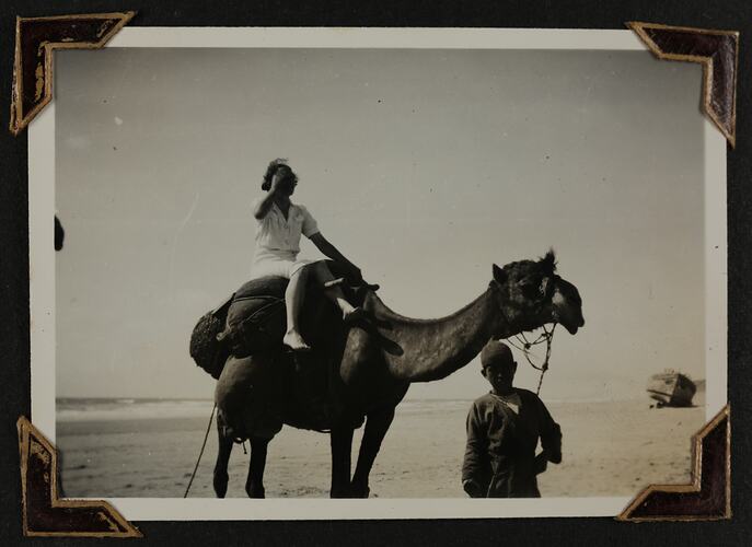 Woman on camelback with boy holding the bridle, beach in background.