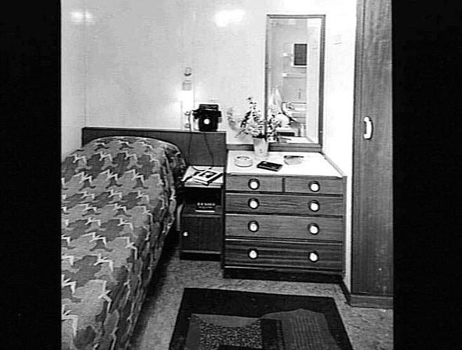 Ship interior. Single bed against left wall. Chest of drawers and stool at right.