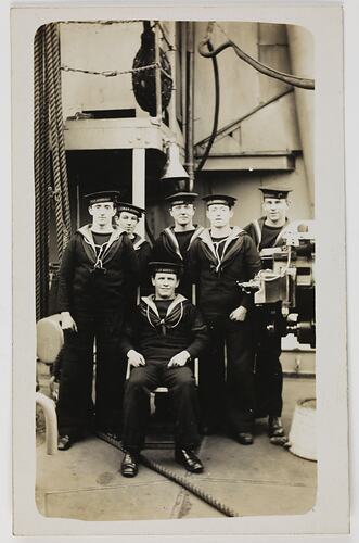Six Navy servicemen on the deck of ship, five standing and one seated in front.
