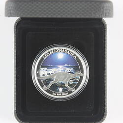 Silver coin with dinosaur. Has dark background. Housed in dark box.