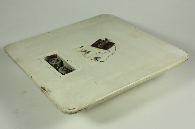 Scuffed white plastic square plate with curved base and rounded corners. Small opening on face side up has two round metal discs within. Above it some wires and a small metal plate are attached to surface.