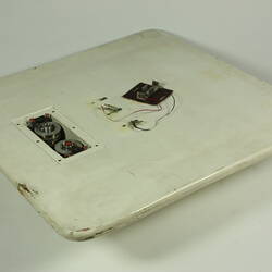 Scuffed white plastic square plate with curved base and rounded corners. Small opening on face side up has two round metal discs within. Above it some wires and a small metal plate are attached to surface.