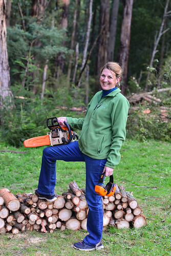 Woman standing near a wood pile holding a chainsaw