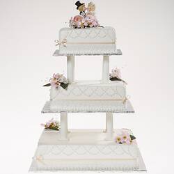 White three tier cake model with pastel ribbons and flowers. Kissing cartoon-style bride and groom on top.