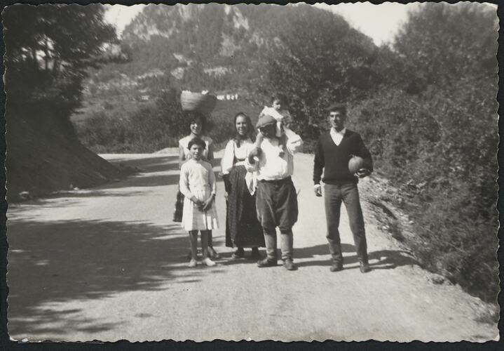 Two women, two children and two men pose on dirt pathway, trees either side. Steep hill behind them.