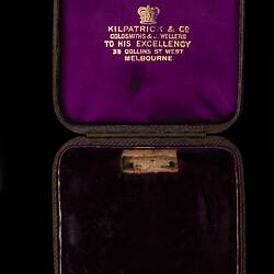 Open jewellery case, purple lined lid with gold lettering. Dark cloth base.