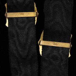 Detail of Back of two black ribbed straps, each with a central gold shield-shaped sliding attachment. Only back of gold sliding bars is visible.