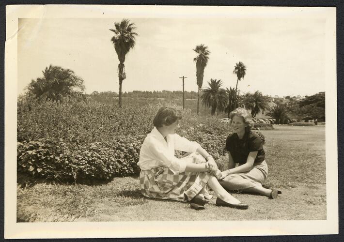 Two women sitting on grass with parkland and gardens behind them.