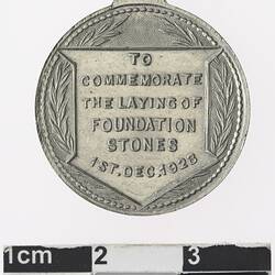 Round silver coloured medal with text surrounded by wreath.
