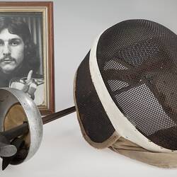 Fencing equipment including epee and mask. Behind them a framed photograph of a man.