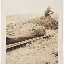 Wrapped up body on a stretcher on the ground, man sitting on a pile of dirt to the right.
