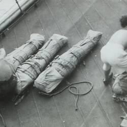 Four sailors preparing and wrapping four bodies wrapped in cloth, rope and belts.