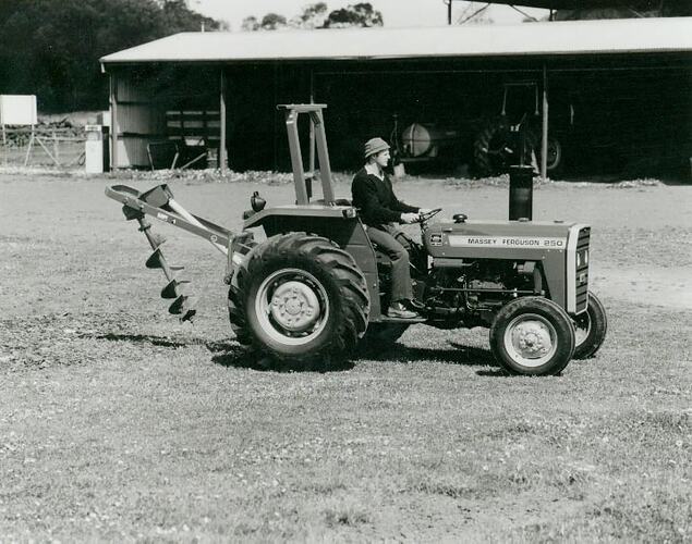 Man driving a  tractor fitted with a Post Hole Digger in front of a farm tractor shed.