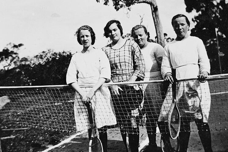 Four girls with tennis racquets, standing at a tennis net.