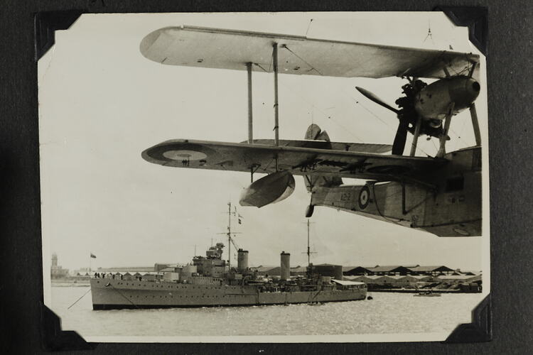 Back half of aircraft in flight with military ship and land in background.