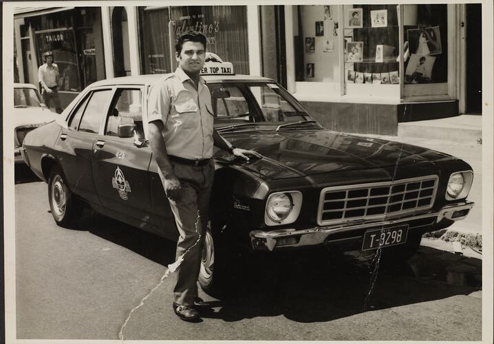 Man in taxi uniform stands beside his taxi parked on a street.