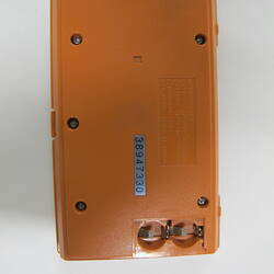 Back of orange plastic, handheld game console, rectangular, closed. Hinge at right side. Battery cover missing