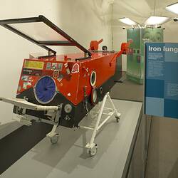 Red rectangular metal box with round opening and lever door at end. On white wheeled stand.