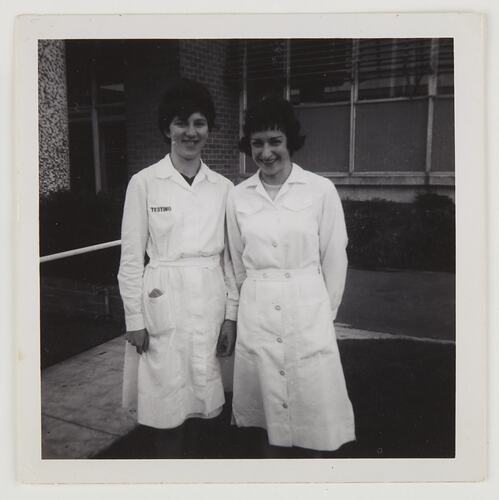 Two women wearing white coat dresses stand outside a factory.