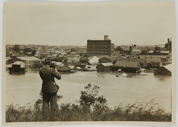 Man taking photograph of factories and other buildings while the Yarra River is in flood in the foreground.