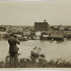 Man taking photograph of factories and other buildings while the Yarra River is in flood in the foreground.
