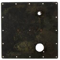 Black metal square (fire-damaged) with holes around edges and two larger ones within. Grey metal rectangular in centre.