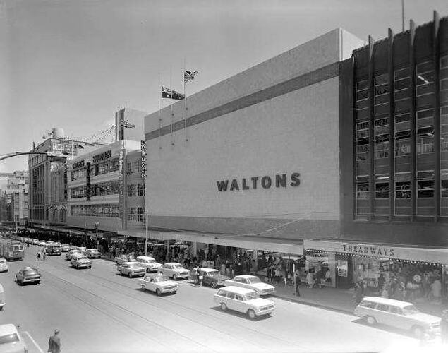 White store with large white front and a WALTONS sign. People crowded at front doors as cars drive past on nea