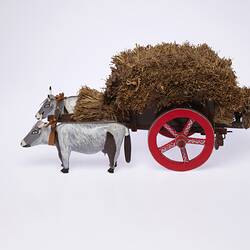 Model of two grey bullocks pulling a hay wagon with haystack and red wheel. Profile view.