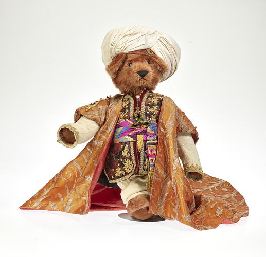 Light brown plush bear wearing white turban, ornate tunic and coat with gold embroidery.