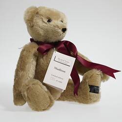 Light brown plush bear with red ribbon around neck with white card attached.