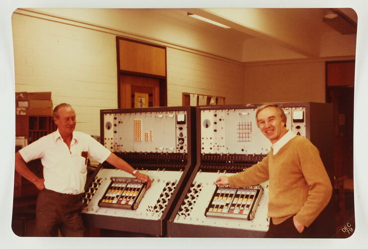 Two men pose in front of two large computers.