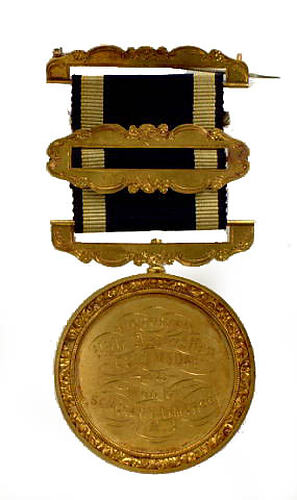 Round gold coloured medal with engraved text, black and gold ribbon attached.