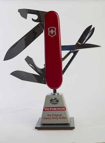 Model of muti-tool on stand. Red handle with all tools out. Large blade, short blade, scissors, bottle opener (with flathead screwdriver), a can opener (with flathead screwdriver), and a reamer.