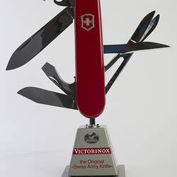 Model of muti-tool on stand. Red handle with all tools out. Large blade, short blade, scissors, bottle opener (with flathead screwdriver), a can opener (with flathead screwdriver), and a reamer.