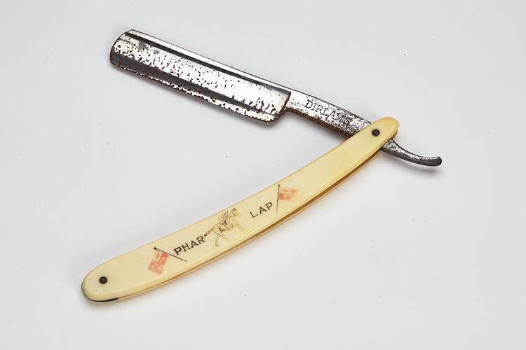 Cut throat razor, metal blade, ivory-styled plastic handle with 'PHAR LAP', picture of jockey on racehorse and two flags. Open.