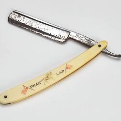 Cut throat razor, metal blade, ivory-styled plastic handle with 'PHAR LAP', picture of jockey on racehorse and two flags. Open.