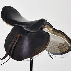 Horse racing saddle. Padded dark brown leather. Double layer beneath saddle flap. Two leather straps on either side. Three quarter front view.