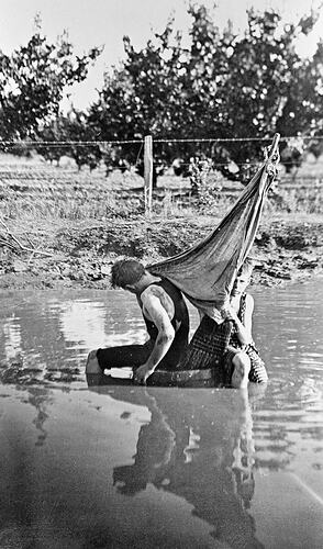 Two boys playing in a water hole with a barrell and sail.