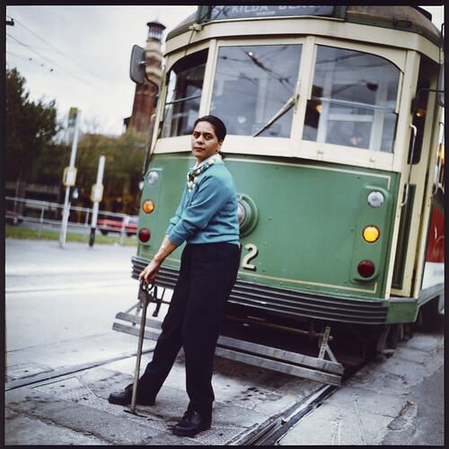 Woman in uniform stands on tram tracks in front of green and yellow tram. She adjusts tracks with a metal bar.