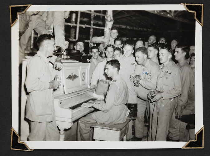 Group of men standing around man playing piano.