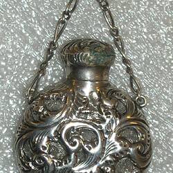 Circular bottle with scrollwork decoration, suspended from chain