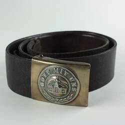 Leather belt with buckle featuring a crown and inscription, belt rolled into circle.