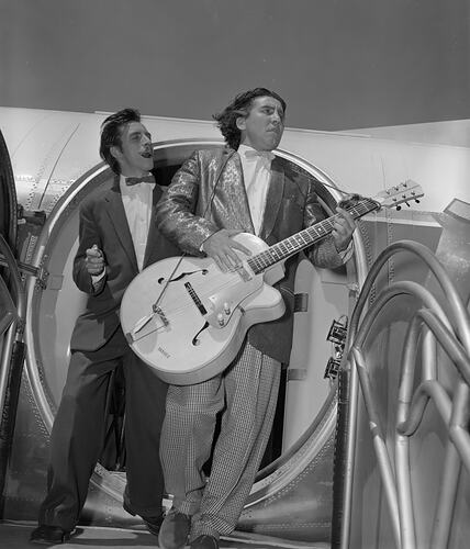 Two men, one holding a large guitar, exiting an aeroplane.